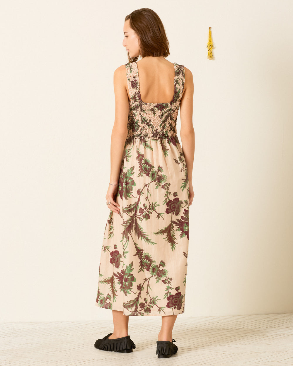 A woman stands indoors against a light-colored wall with a small tassel decoration on the right, wearing the Kay Summermoss Dress—a sleeveless, backless cotton-silk midi dress featuring a smocked bodice and botanical motif—paired with black shoes.