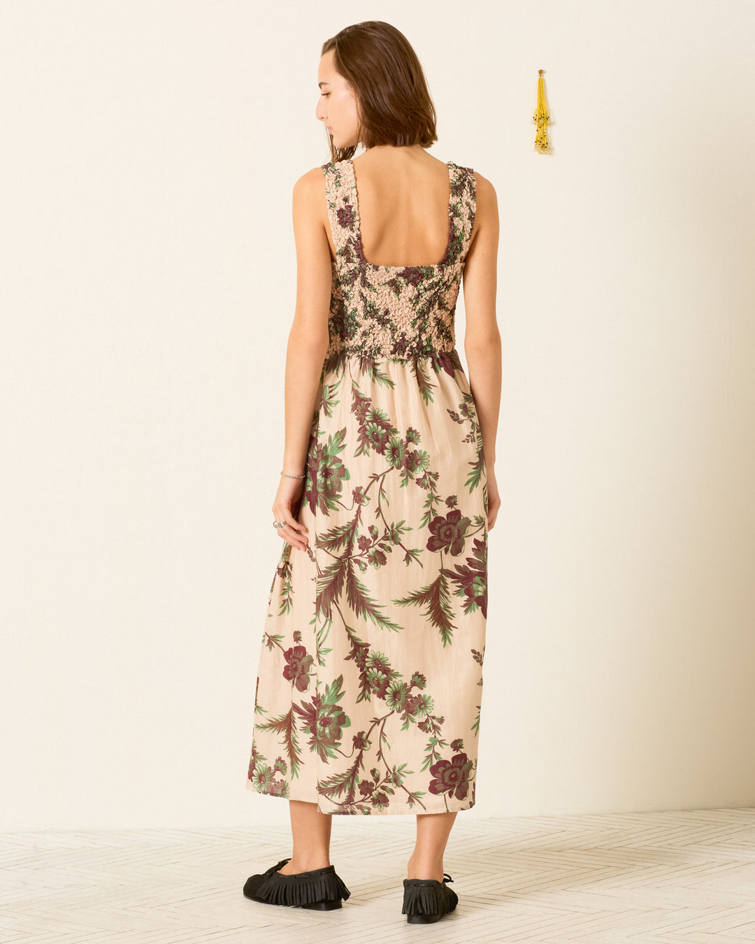 A woman stands indoors against a light-colored wall with a small tassel decoration on the right, wearing the Kay Summermoss Dress—a sleeveless, backless cotton-silk midi dress featuring a smocked bodice and botanical motif—paired with black shoes.