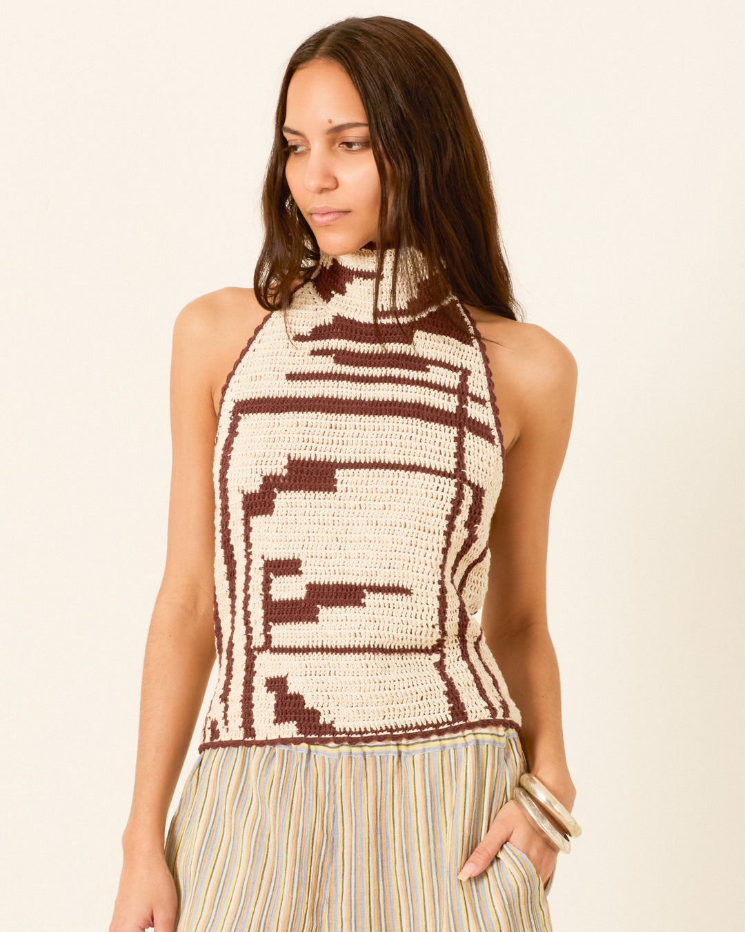 A woman wears the Knox Auburn Driftmap Top, a sleeveless high-neck knit with brown and cream ikat-inspired geometric patterns, styled with striped pants and silver bangles.