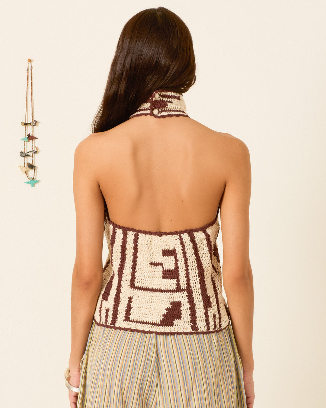 A person with long brown hair is shown from the back, wearing the Knox Auburn Driftmap Top—a crocheted halter with a geometric ikat-inspired pattern—paired with a pleated skirt.