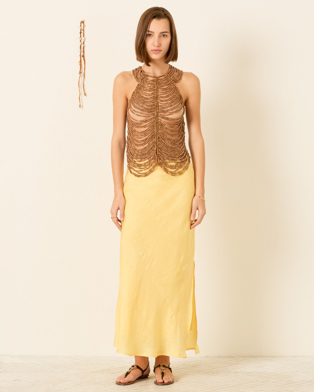 A woman faces forward in the Lania Fawn Wood Top, paired with a long yellow skirt and sandals, against a plain light background.