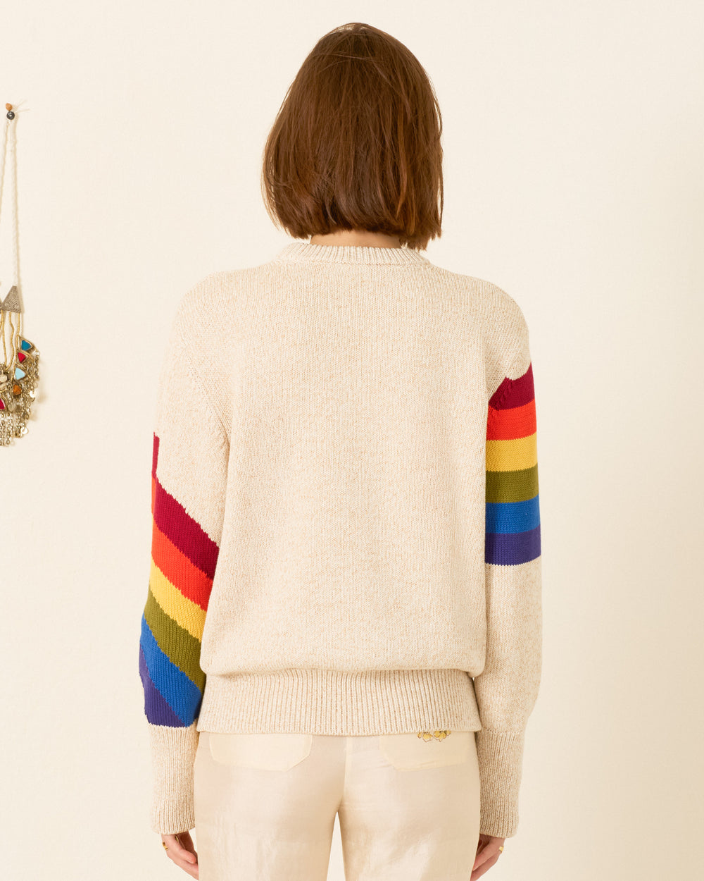 A person stands facing away, wearing the Sammi Prism Knit Cardigan—beige, with rainbow motif sleeves—paired with light-colored pants, crafted from organic Italian cotton.