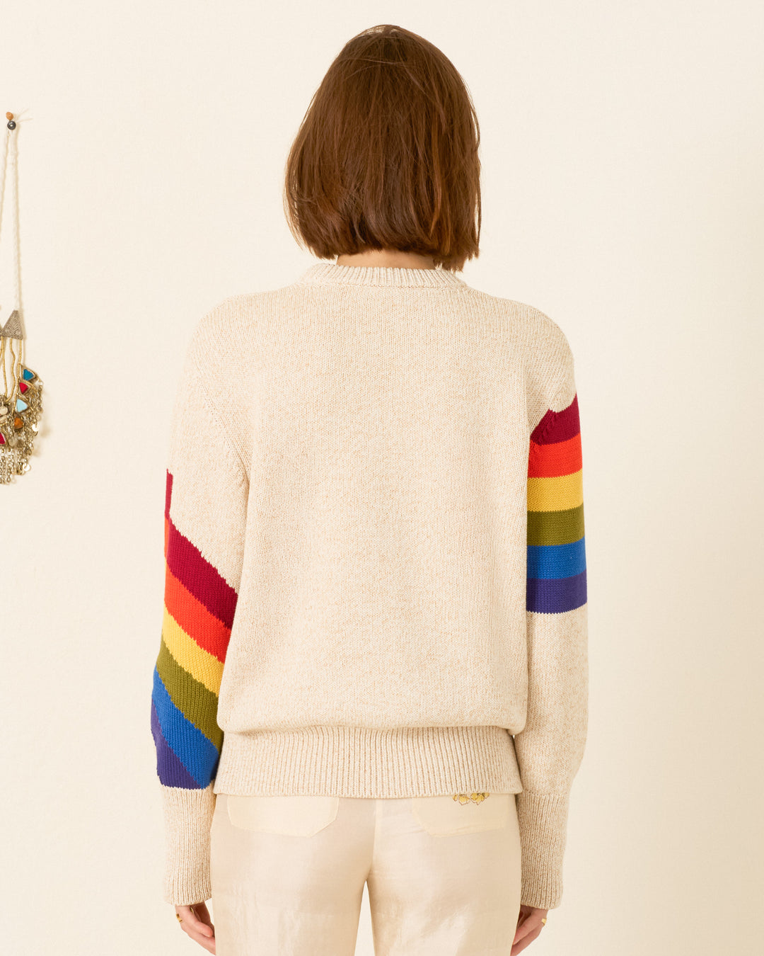 A person stands facing away, wearing the Sammi Prism Knit Cardigan—beige, with rainbow motif sleeves—paired with light-colored pants, crafted from organic Italian cotton.
