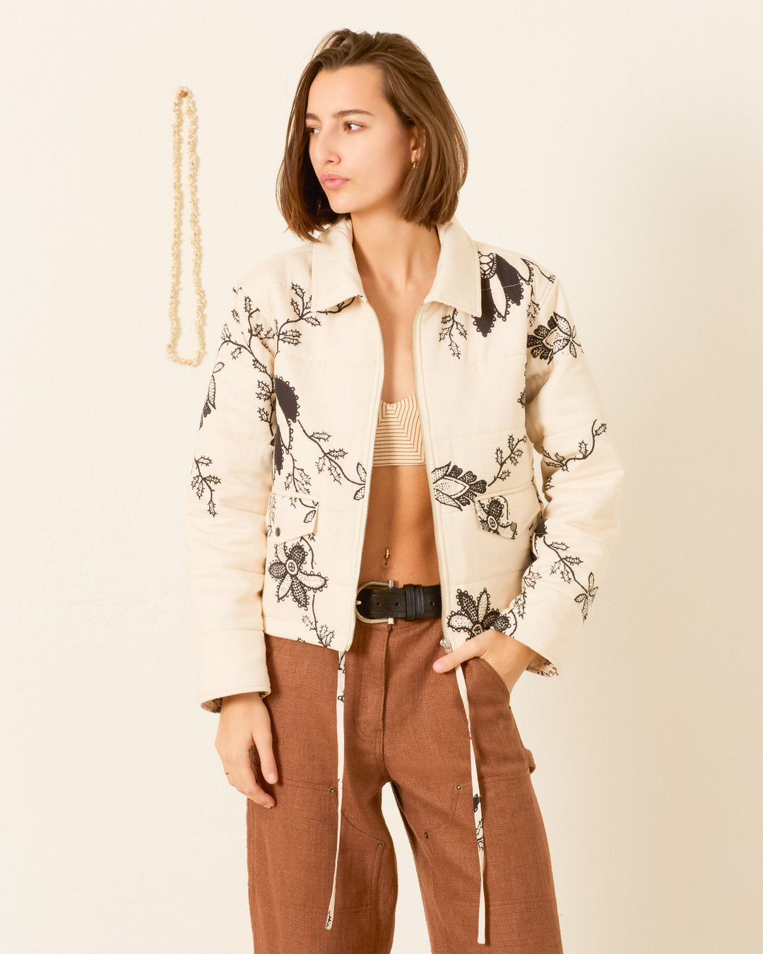 Person wearing the Lex Shadow Lace Jacket—a light-colored, floral embroidered workwear-inspired piece—paired with a striped bandeau top and brown pants, standing against a plain wall with a beaded decoration hanging beside them.