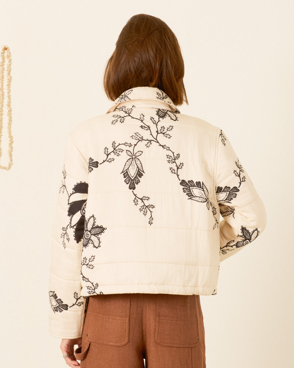 A person wearing the Lex Shadow Lace Jacket—a cream-colored, workwear-inspired quilted jacket with black floral embroidery—stands facing away to show the back of the jacket and brown pants.