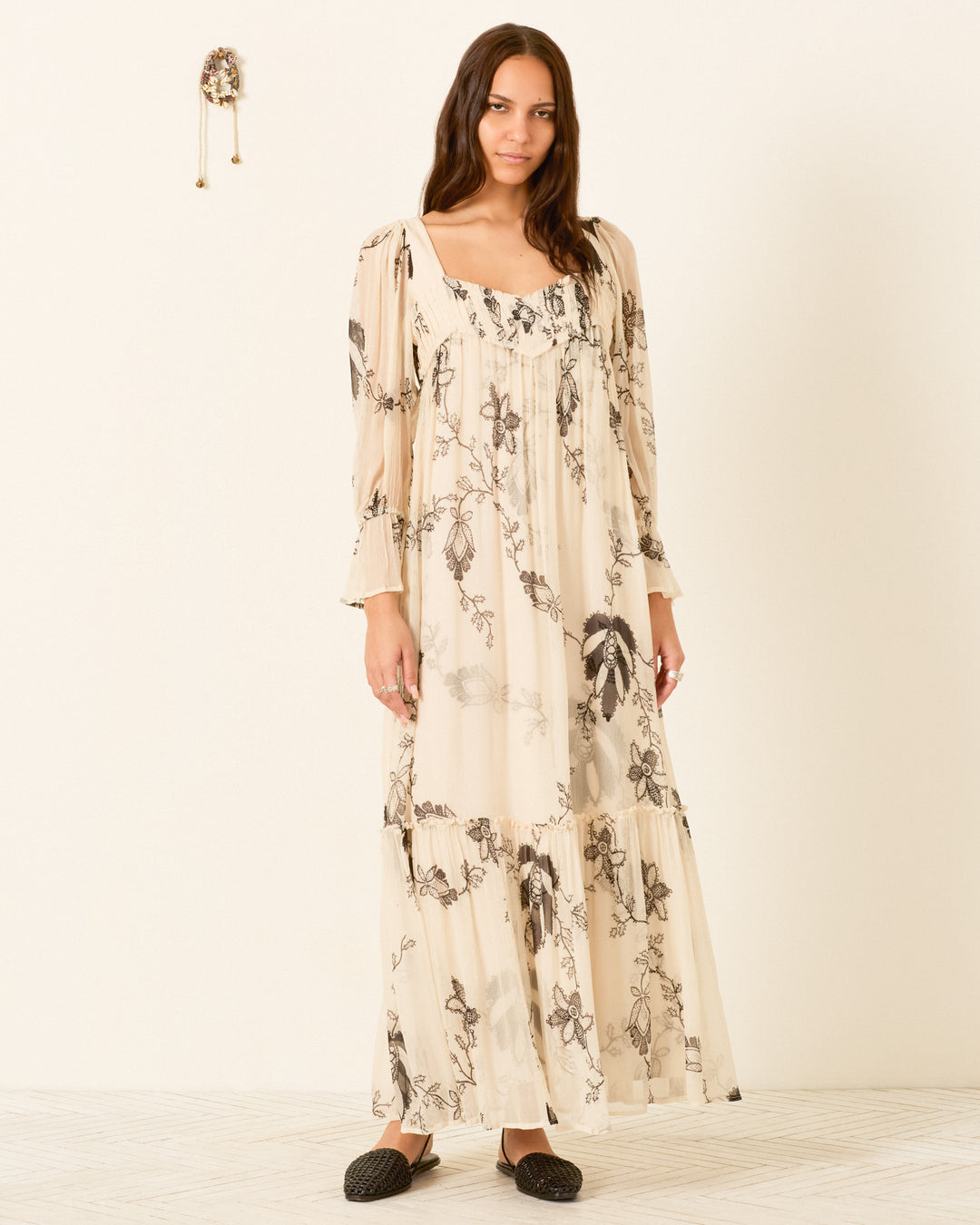 A woman stands indoors against a plain wall, wearing the Lilium Shadow Lace Dress—a long, cream-hued floral maxi featuring puff sleeves and a pleated empire waist—paired with black slip-on shoes.