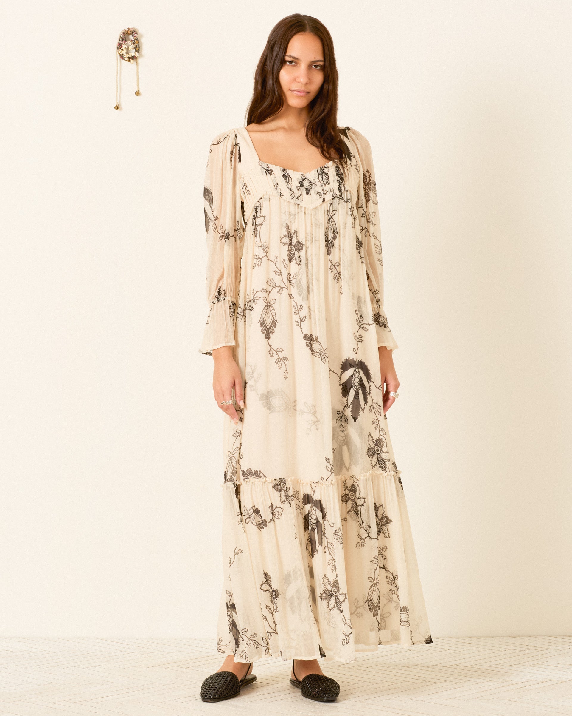 A woman stands indoors against a plain wall, wearing the Lilium Shadow Lace Dress—a long, cream-hued floral maxi featuring puff sleeves and a pleated empire waist—paired with black slip-on shoes.