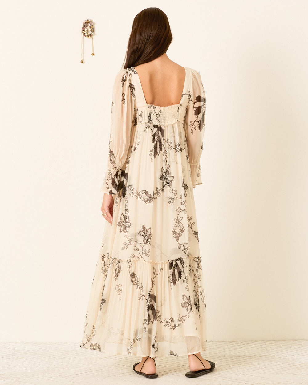 Woman with long brown hair wearing the Lilium Shadow Lace Dress—a cream, floral-patterned, long-sleeve maxi dress with puff sleeves—paired with black sandals, standing indoors and facing away from the camera.