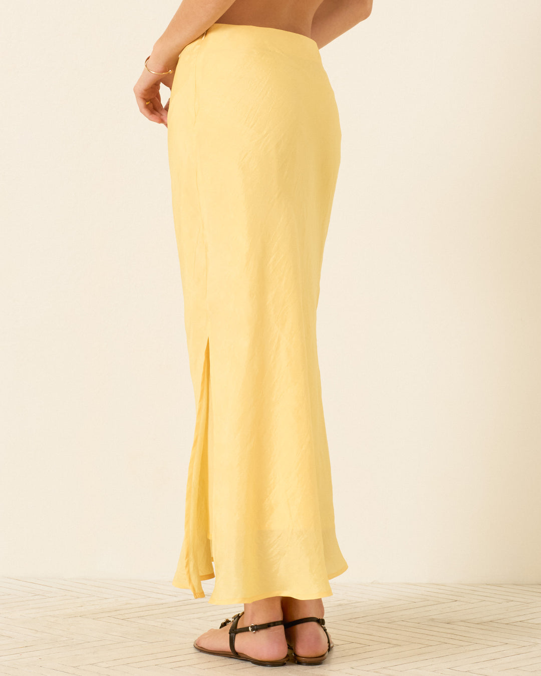 A person wears the Lyra Sunlight Silk Skirt, a long yellow artisan-made skirt with a side slit, paired with black sandals, standing on a light-colored floor against a plain background.