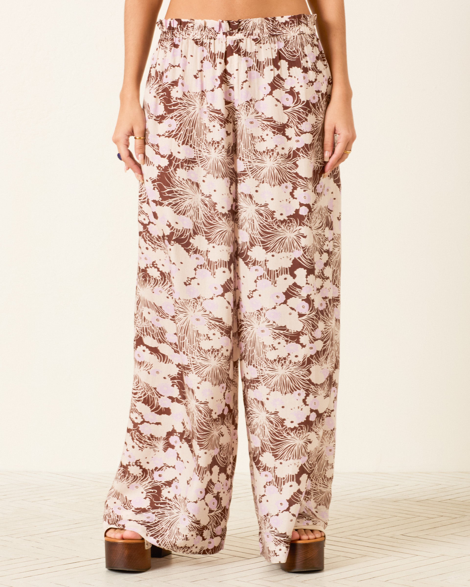 Wearing the Margie Dandelion Dusk Pant, featuring a high-waisted, wide-leg silhouette and a delicate floral pattern in brown and beige hues, paired with platform sandals against a light background.