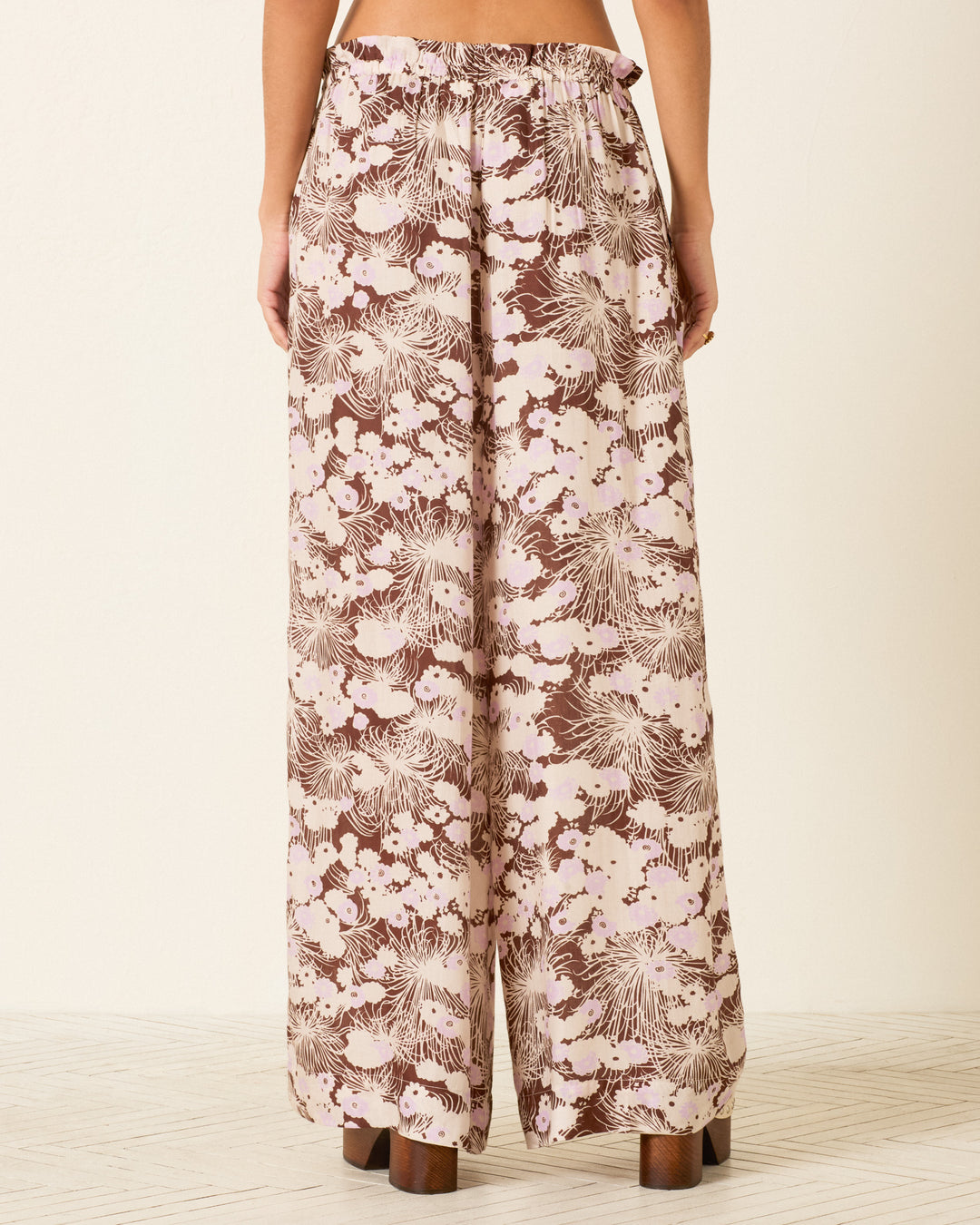 A person wearing the Margie Dandelion Dusk Pant—a pair of high-waisted, wide-leg Tencel pants featuring a brown and cream floral motif—is viewed from the back while standing on a light-colored floor.