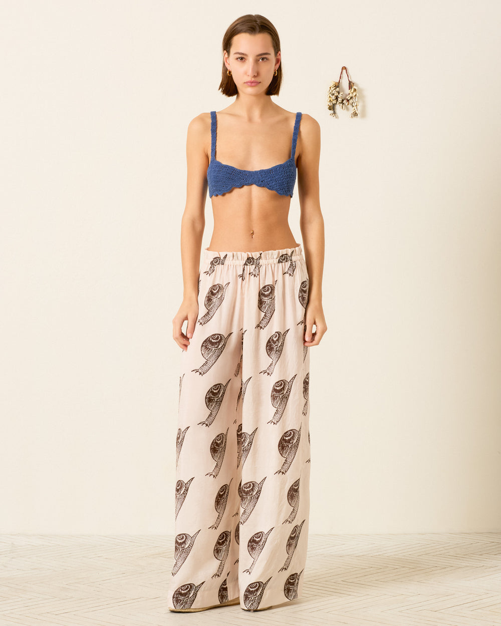 A woman stands indoors wearing the Margie Snail Study Silk Pant, featuring a wide-leg silhouette and a hand-block-printed snail design. Dried flowers hang on the wall behind her.