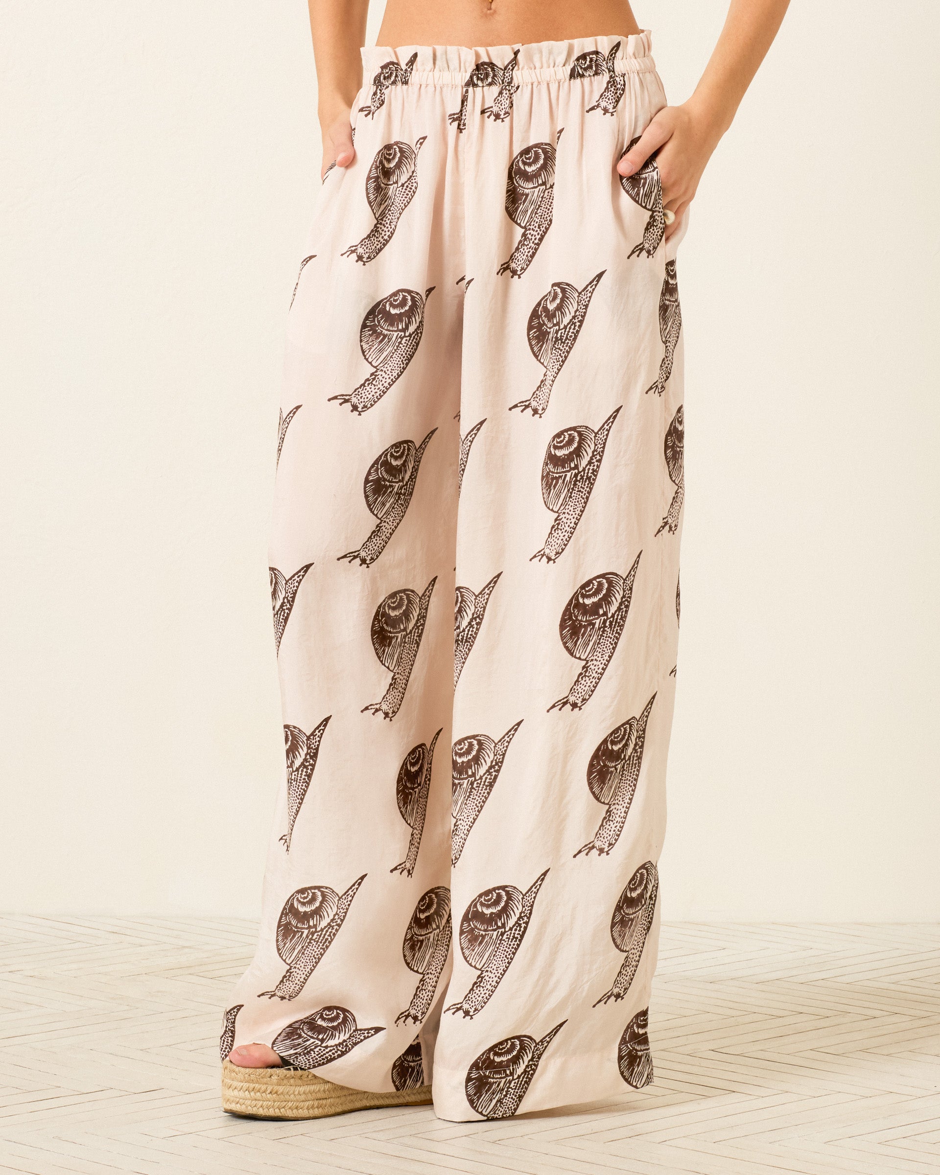 Wearing Margie Snail Study Silk Pant—light-colored, hand-block-printed wide-leg pants with all-over snail print. Lower body visible; standing on a patterned floor, hands in pockets.