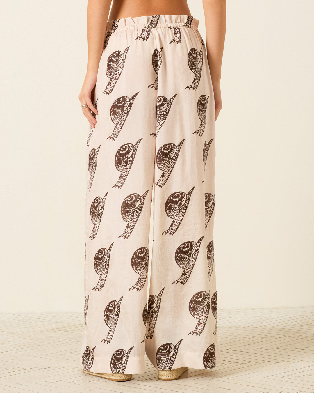 A person wearing Margie Snail Study Silk Pant, featuring wide legs and a hand-block-printed all-over black fish design, is photographed from the back.