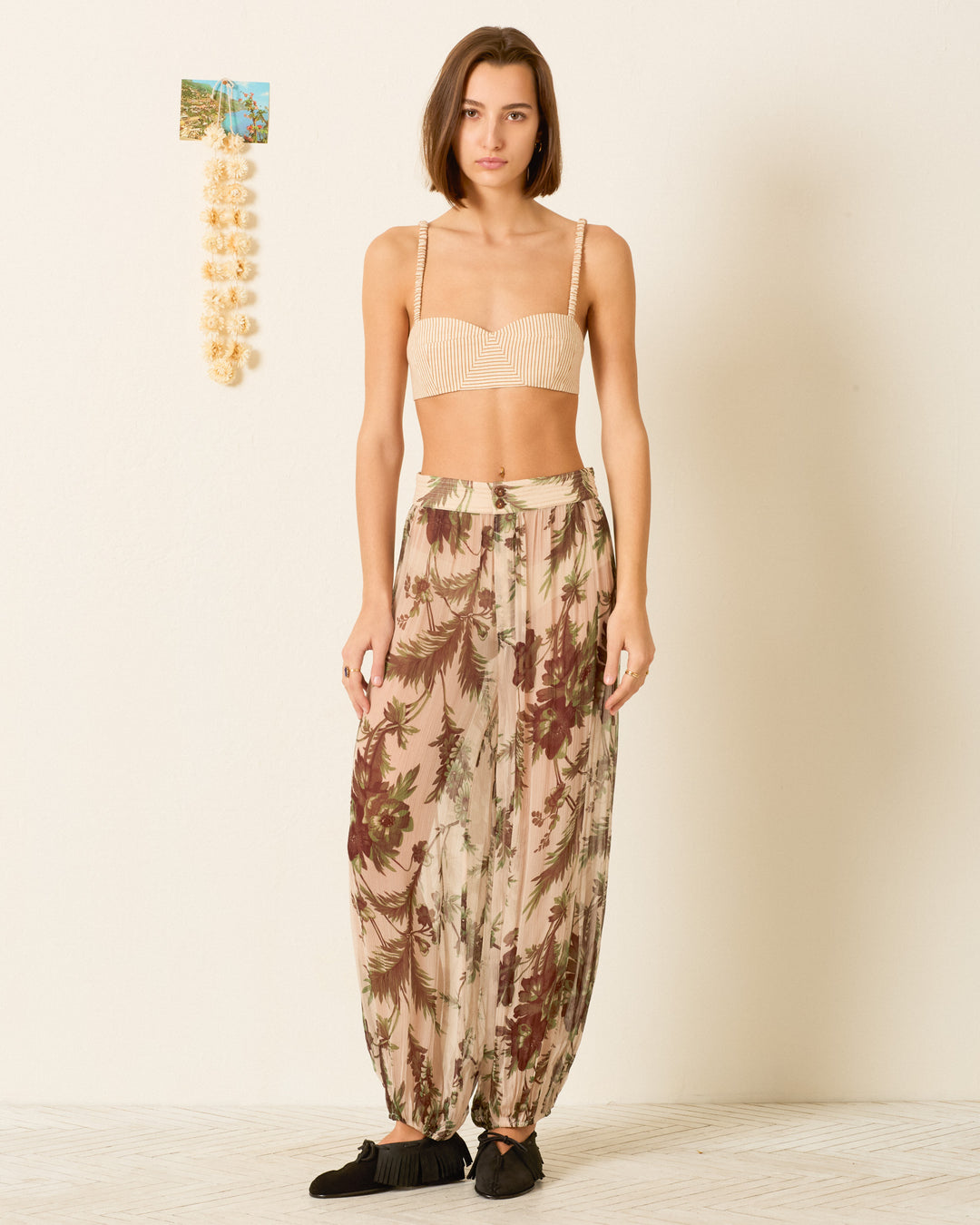 A woman stands indoors wearing a beige crop top and the Margot Summermoss Pant—loose, floral, harem-style—paired with black shoes. The background has a small photo and a hanging string of shells on the wall.