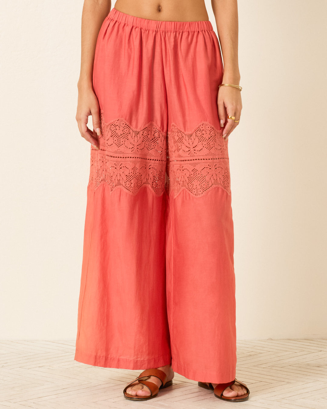 A person wearing the Margie Rouge Dust Pant, featuring a coral cotton-silk blend with lace panels, paired with a gold bracelet and brown sandals, stands against a neutral background.