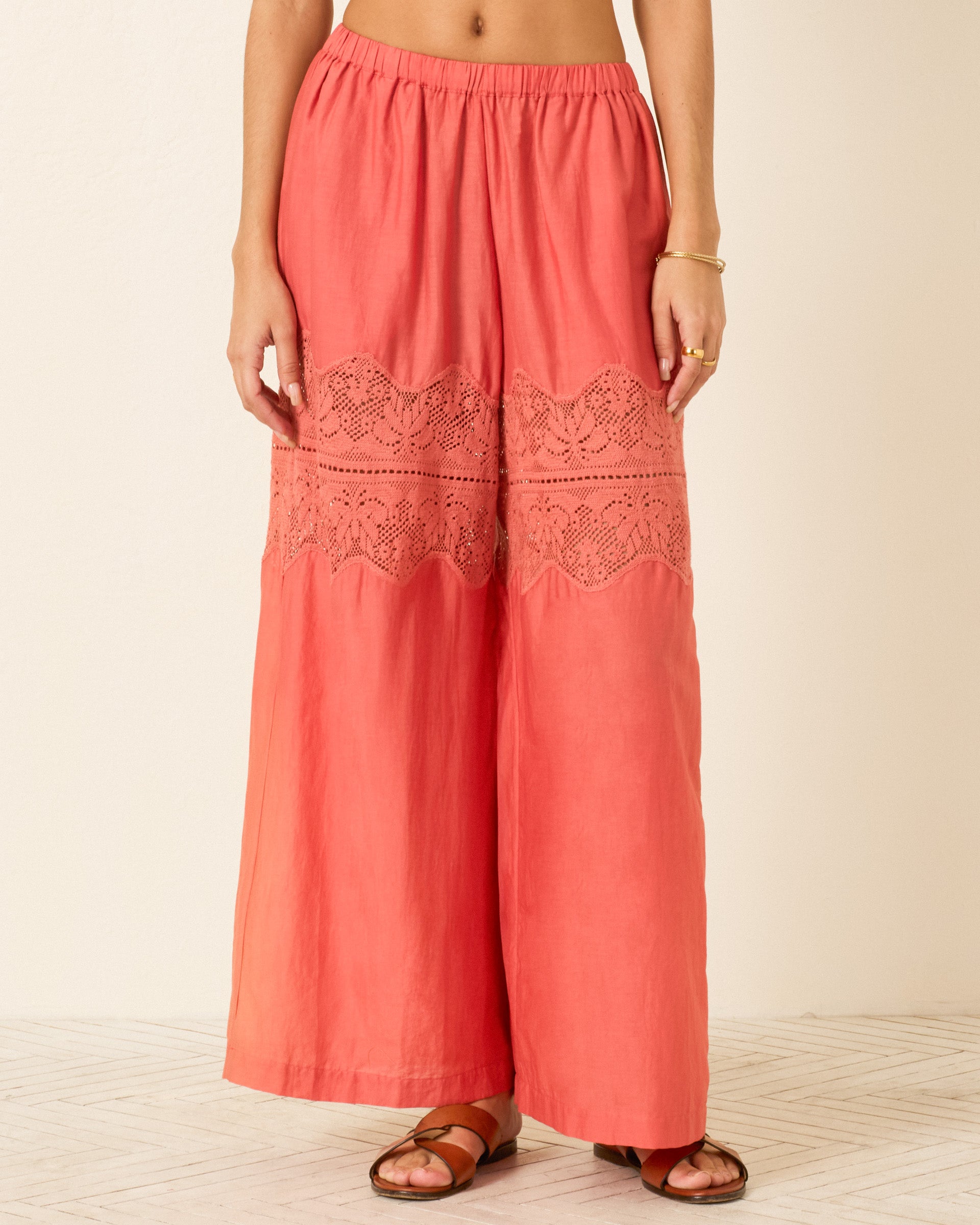 A person wearing the Margie Rouge Dust Pant, featuring a coral cotton-silk blend with lace panels, paired with a gold bracelet and brown sandals, stands against a neutral background.