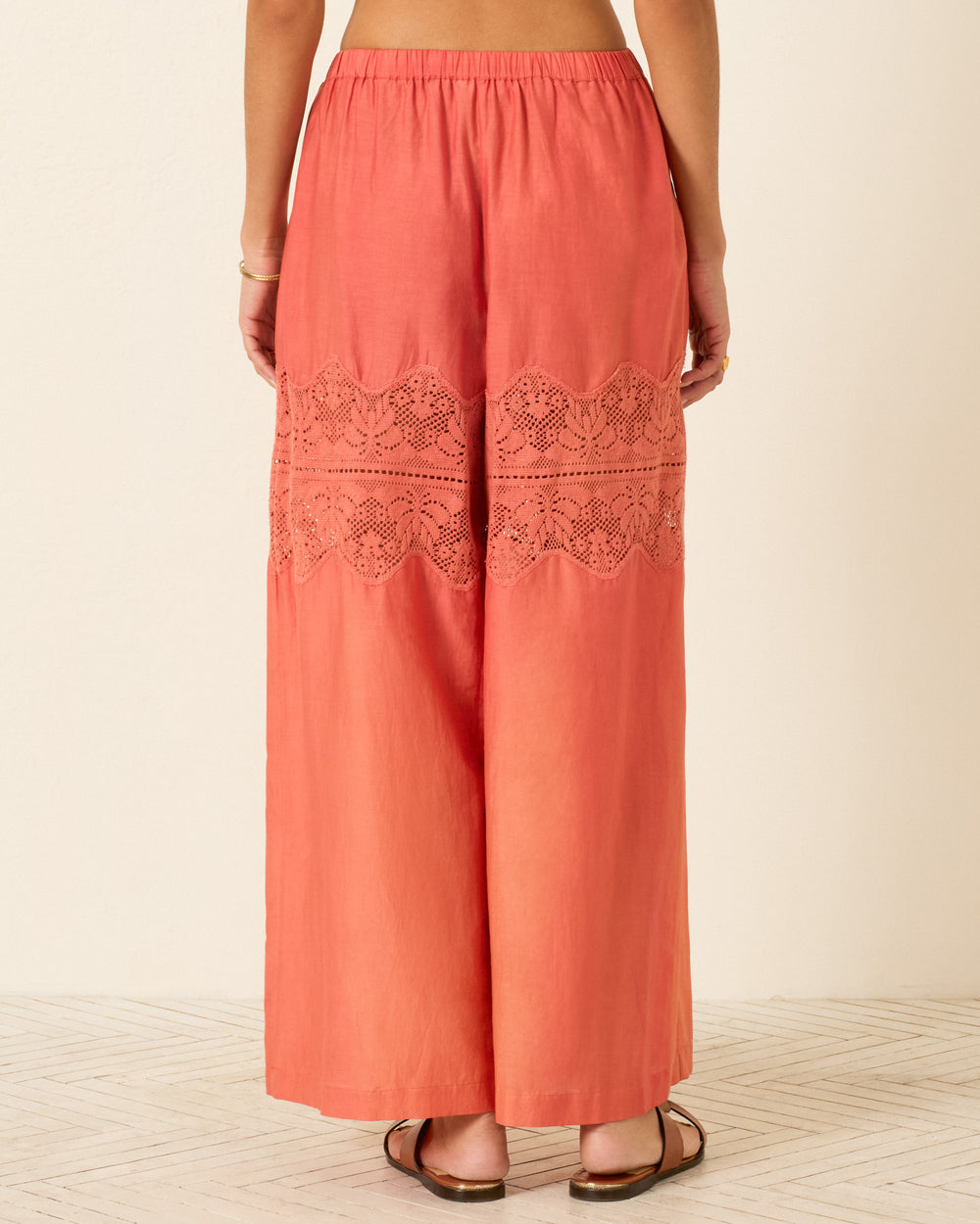 Viewed from the back, a person stands on a light floor wearing the Margie Rouge Dust Pant—coral wide-leg pants made from a cotton-silk blend with delicate cotton lace detailing at the upper calves.
