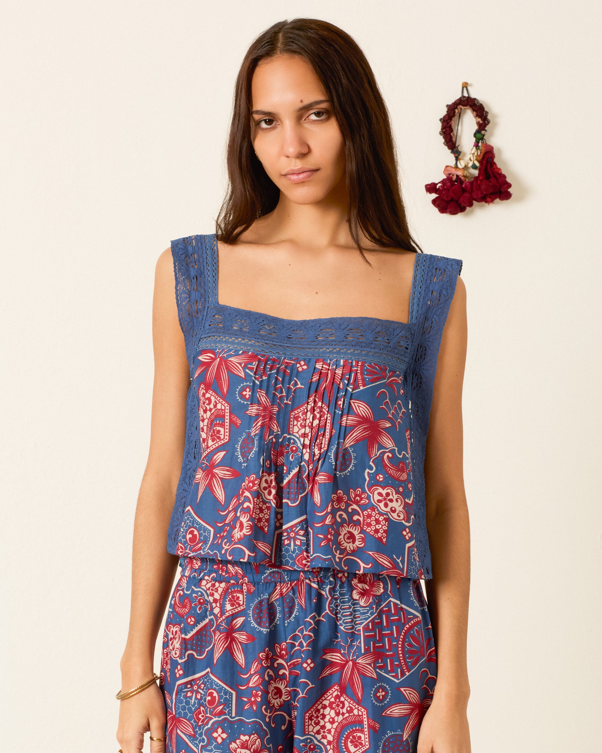 A woman with long brown hair wears the Mimi Delft Hive Top, a blue and red wagara-print cropped top, paired with matching pants while standing indoors near a wall with a decorative hanging.