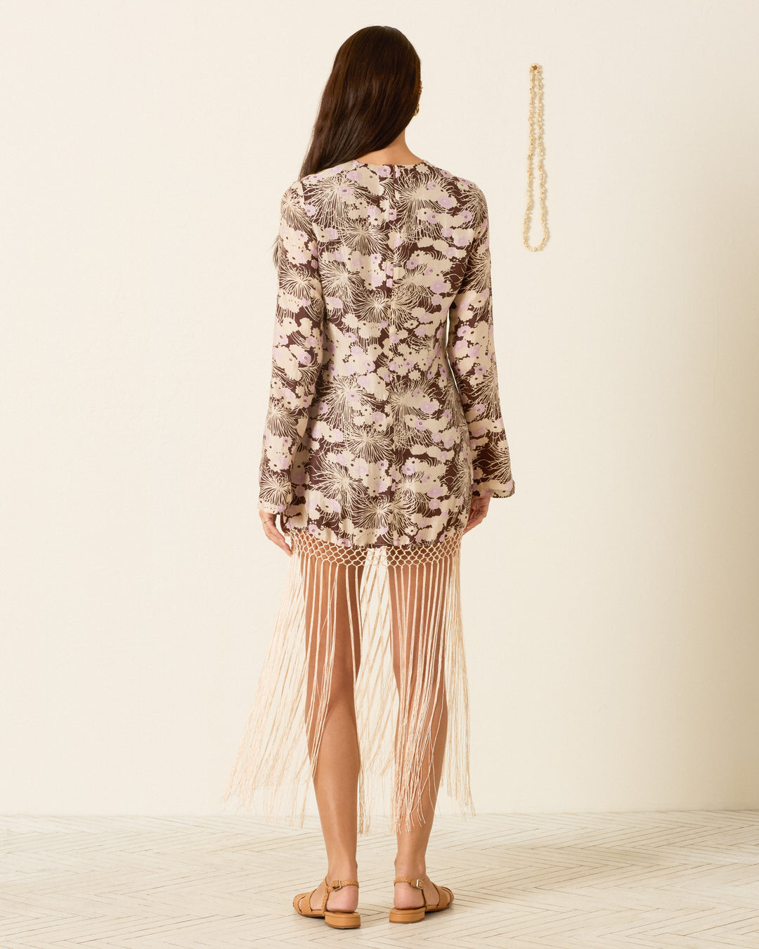 A woman stands with her back to the camera, wearing the Narene Dandelion Silk Dress—featuring a floral motif, mini length, and fringe hem—paired with flat sandals against a plain light-colored wall.