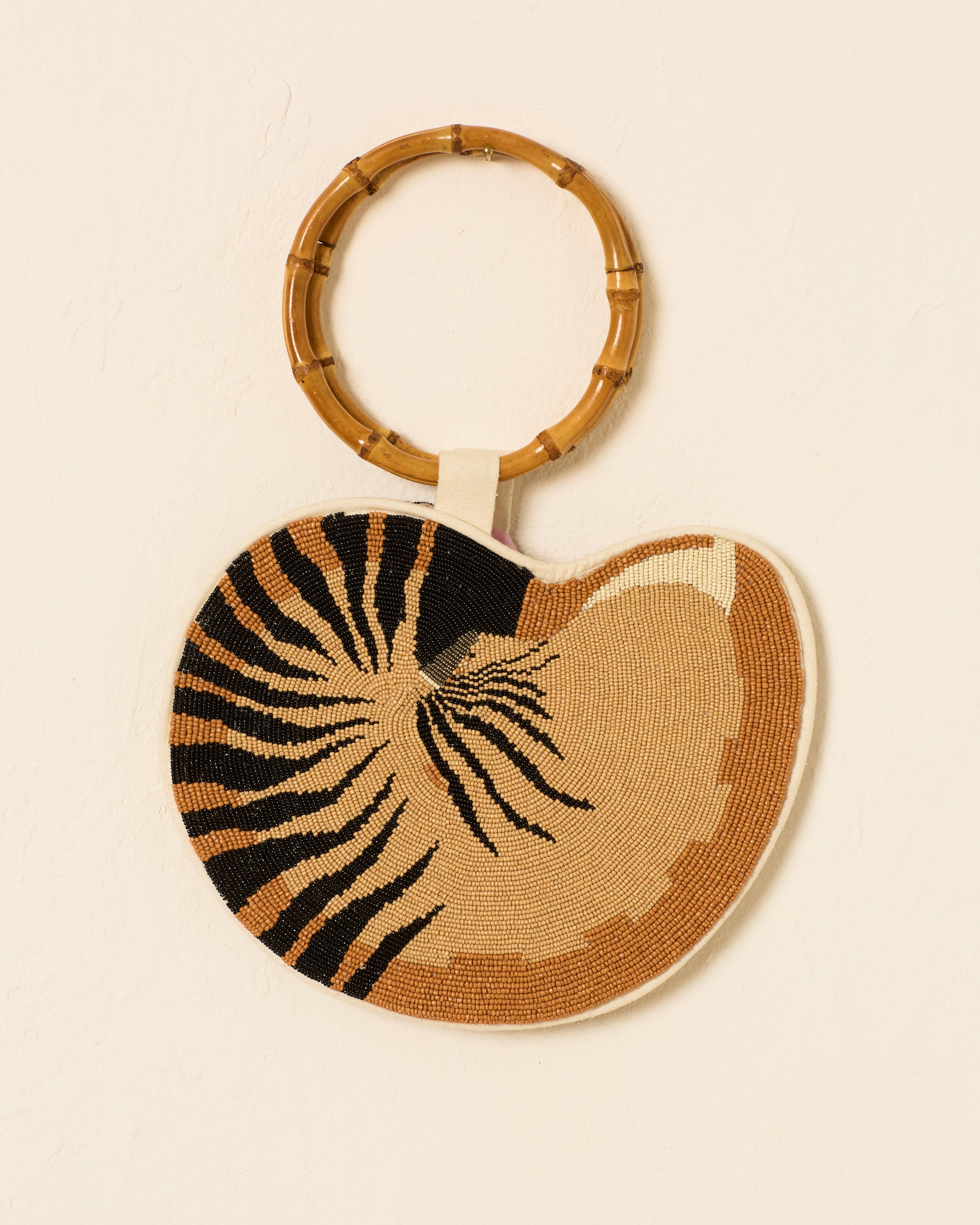 The Nautilus Sandbar Beaded Bag, featuring a seashell-inspired design in brown, tan, and black with a round bamboo handle, hangs against a plain light wall.