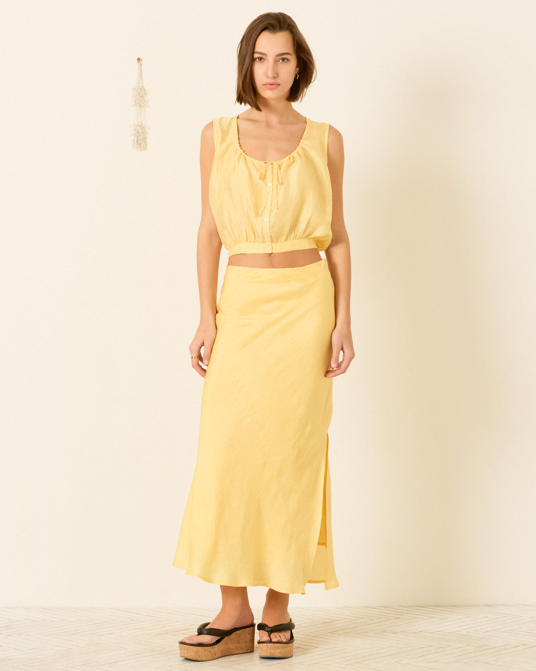 A woman stands indoors wearing the Nova Sunlight Silk Shirt, an artisan-made sleeveless yellow crop top, and a matching long skirt with a side slit, paired with black platform sandals.