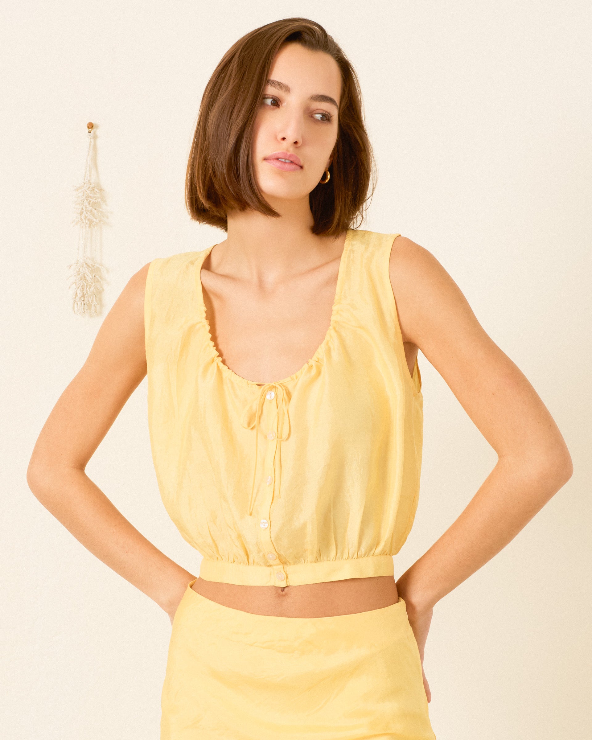 A woman with a bob haircut wears the Nova Sunlight Silk Shirt, an artisan-made sleeveless blouse in light yellow, paired with a matching skirt, standing by a plain white wall with minimal decoration.