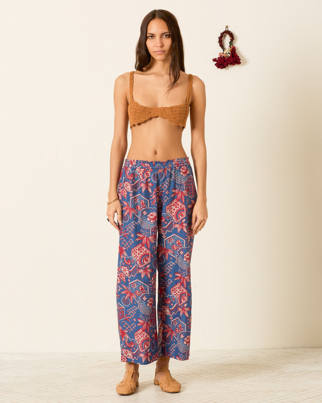 A woman stands indoors by a beige wall, wearing a brown crochet bralette, the blue and red Otto Delft Hive Pant in cotton-silk, and tan shoes. A small wall decoration is visible behind her.