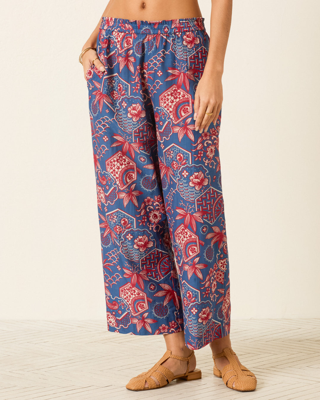 A person wearing the Otto Delft Hive Pant—wide-leg blue cotton-silk pants with a red and white floral geometric wagara print—stands on a light floor with tan woven sandals.