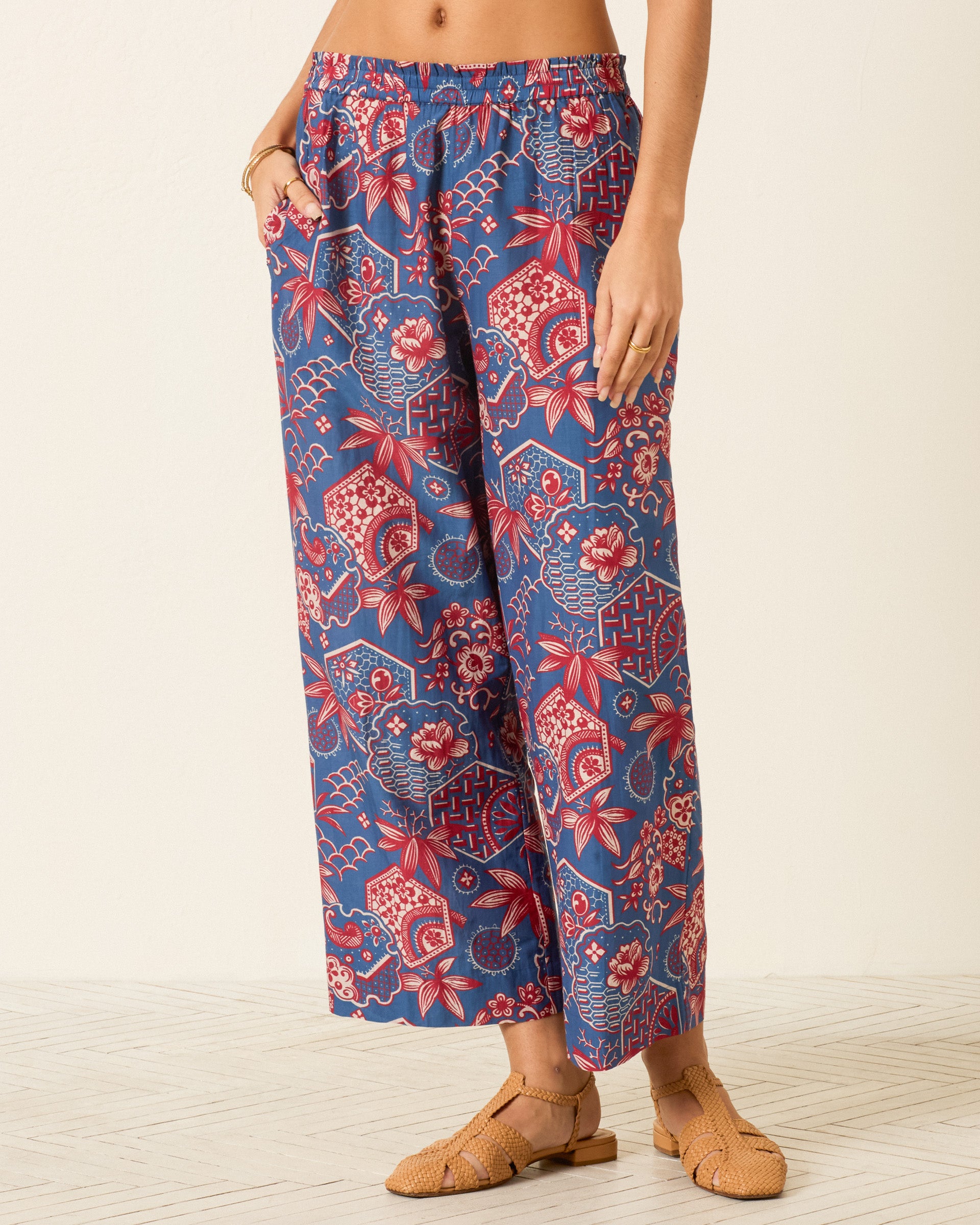 A person wearing the Otto Delft Hive Pant—wide-leg blue cotton-silk pants with a red and white floral geometric wagara print—stands on a light floor with tan woven sandals.