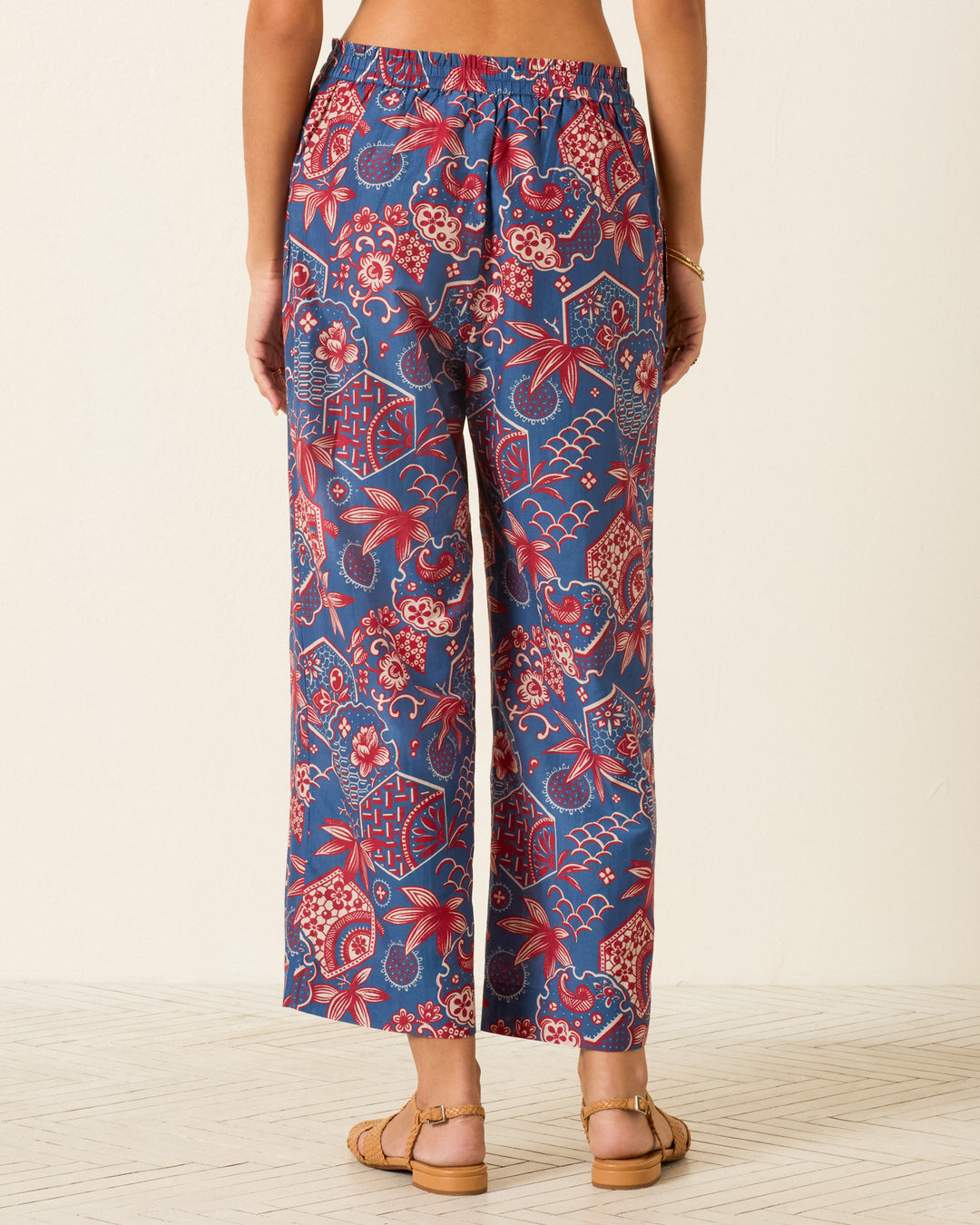 A person wearing the Otto Delft Hive Pant, featuring blue and red wagara print wide-leg cotton-silk design, paired with tan sandals stands on a light floor facing away from the camera.