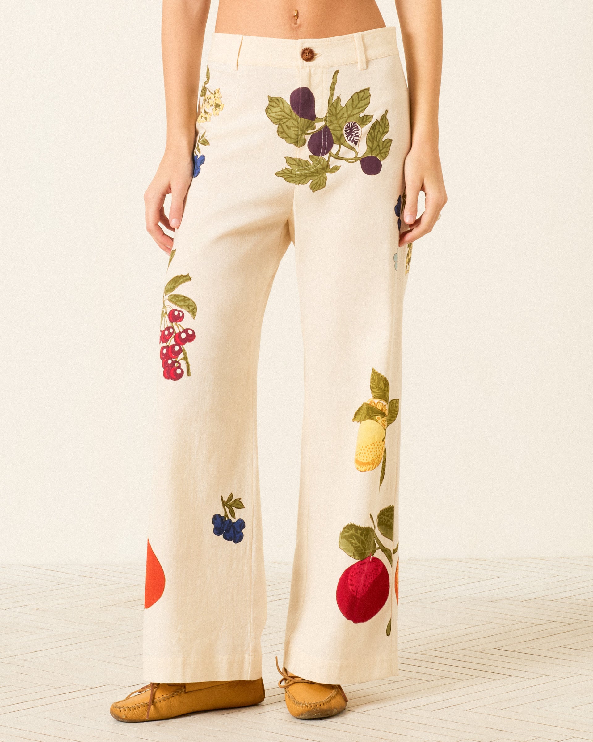 Model wears the Paige Nine Fruit Pant—cream Khadi cotton wide-leg pants with colorful embroidered fruit designs—paired with brown shoes, standing against a neutral background.