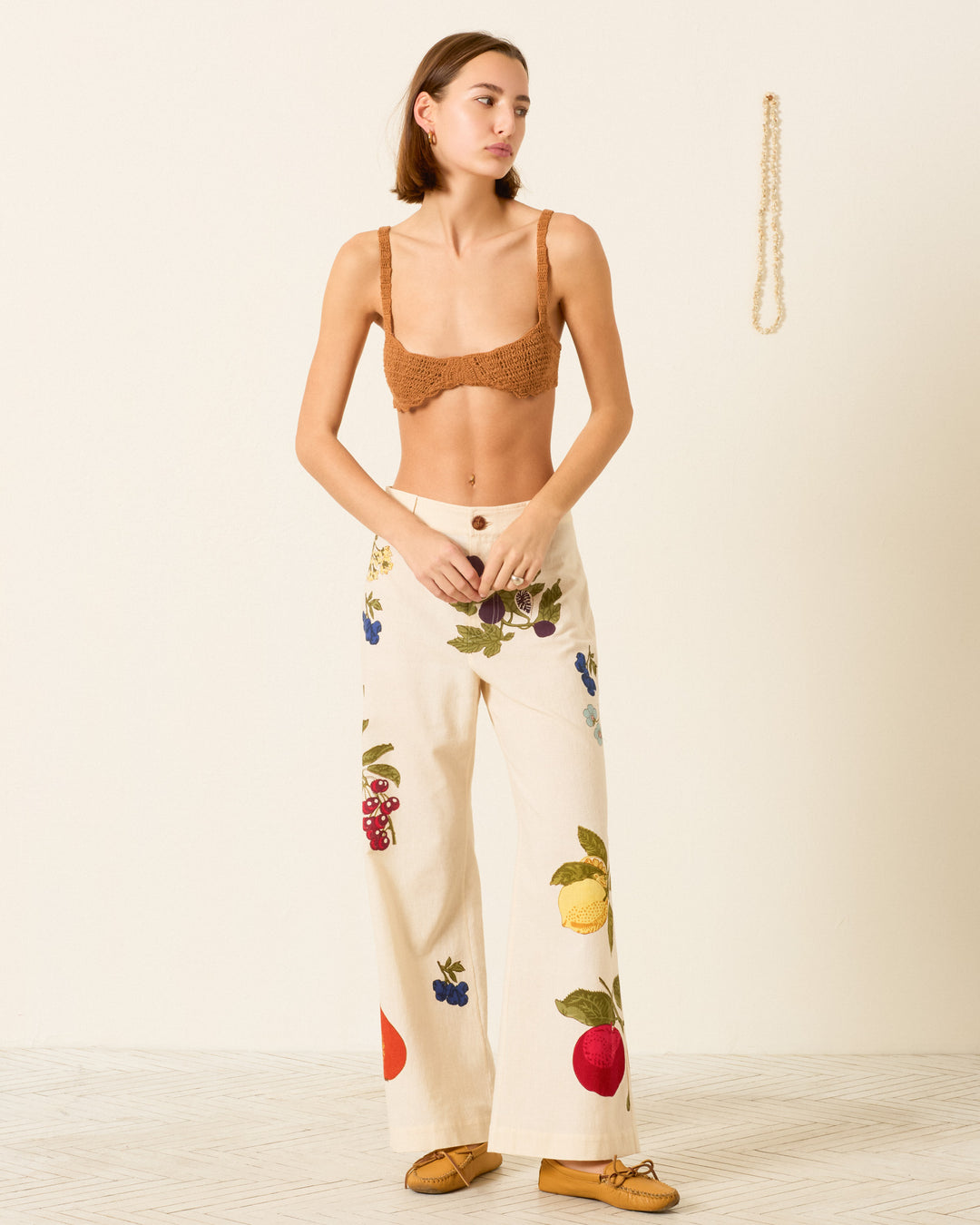 A woman stands indoors wearing a brown crochet bralette, Paige Nine Fruit Pant with hand-block-printed fruit motifs, and brown shoes. A beaded necklace hangs on the wall behind her.