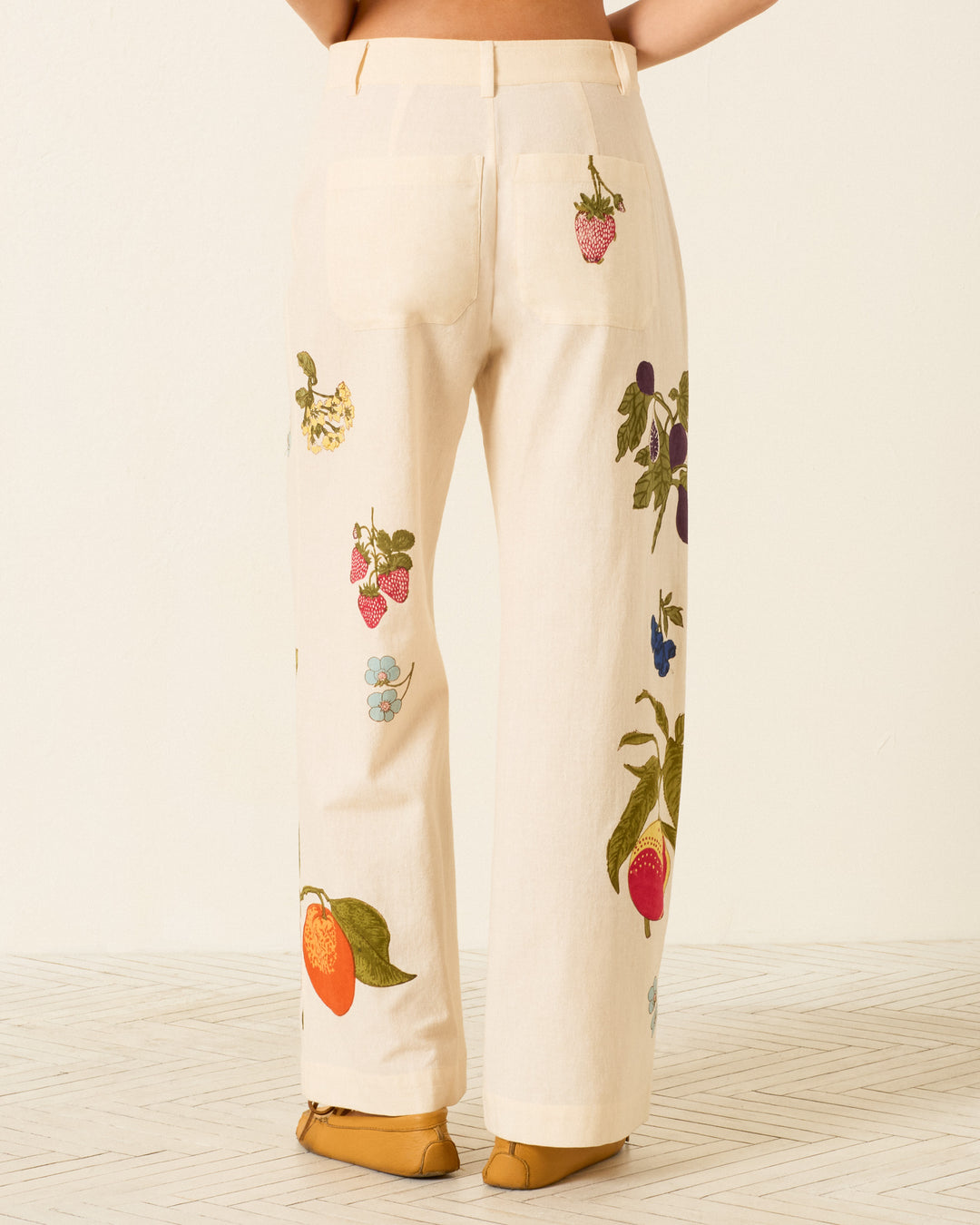 A person wearing Paige Nine Fruit Pant—cropped Khadi cotton pants with colorful fruit embroidery on the legs—stands on a light textured floor, shown from the back.