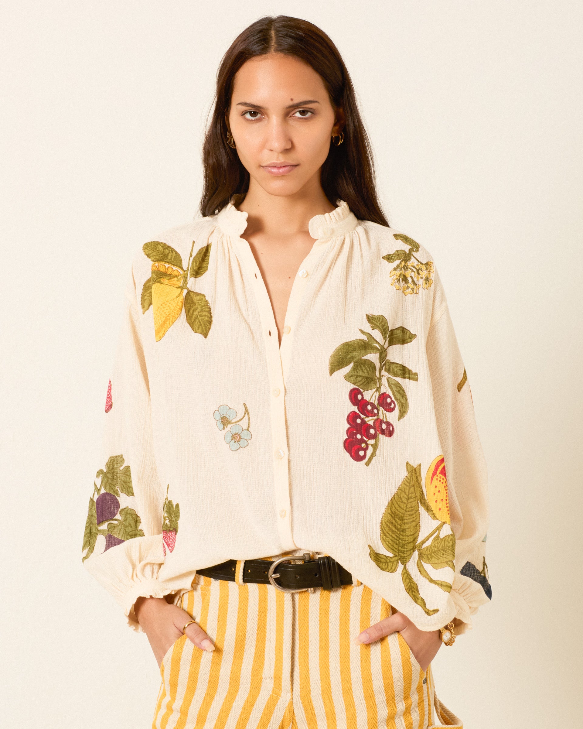 A woman wearing the Poet Nine Fruit Blouse, a cream hand-block-printed top with colorful botanical prints, paired with yellow striped pants, stands with her hands in her pockets against a plain background.