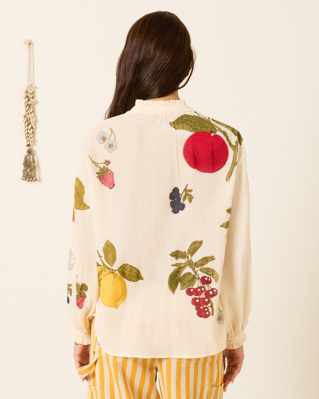 A person with long brown hair faces away from the camera against a plain wall, wearing the Poet Nine Fruit Blouse—a hand-block-printed cream top featuring colorful fruit prints—paired with yellow striped pants.