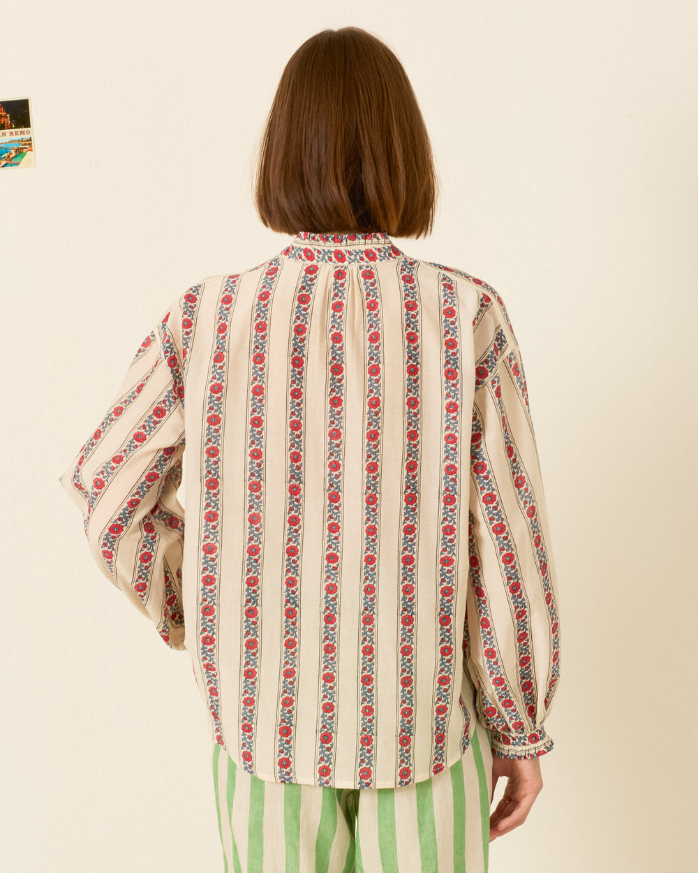 A person with straight, shoulder-length brown hair wears the Poet Roseband Stripe Blouse and green striped pants, standing with their back to the camera against a plain wall.