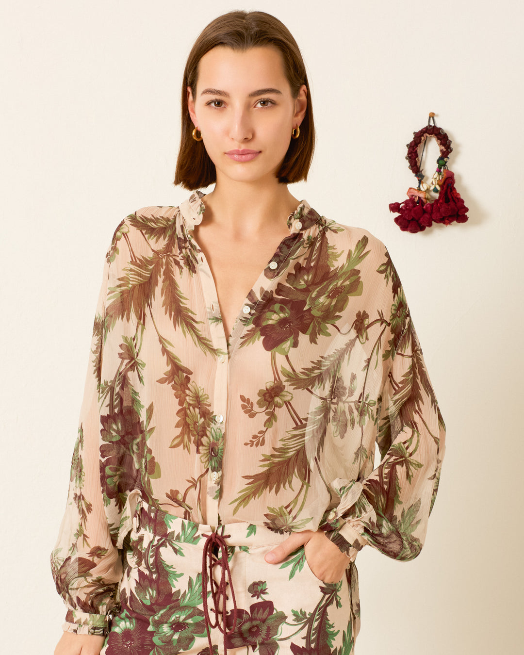 A woman wears the Poet Summermoss Blouse—sheer, long-sleeve cupro-chiffon with a botanical motif—and matching pants, standing against a light wall with a decorative item in the background.