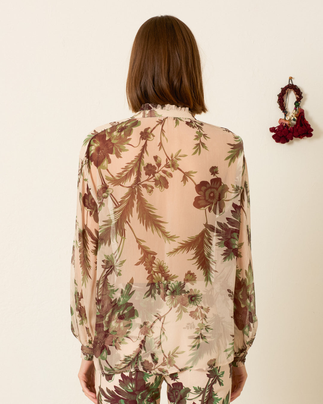 A woman with shoulder-length brown hair, seen from behind, wears the Poet Summermoss Blouse featuring a sheer cupro-chiffon botanical motif in green and burgundy. A small hanger with dark flowers is on the wall to her right.