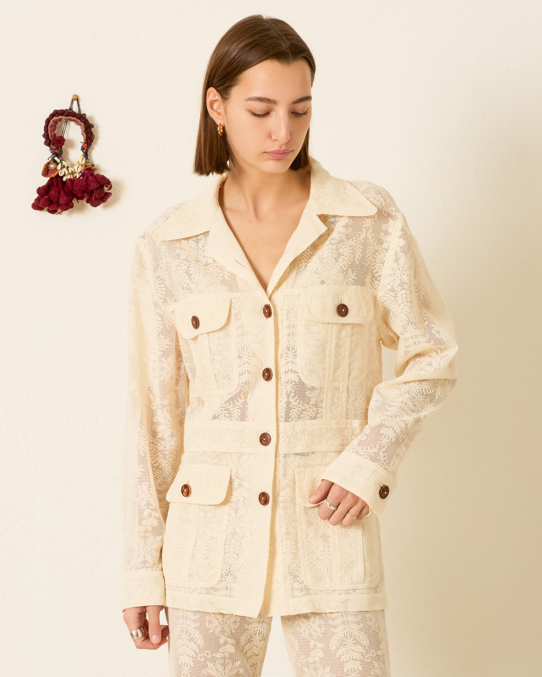 A woman stands against a plain light wall, wearing the Reno Pearl Lace Lattice Jacket—a cream, long-sleeve lace jacket with four front pockets. A small red decorative item hangs on the wall beside her.
