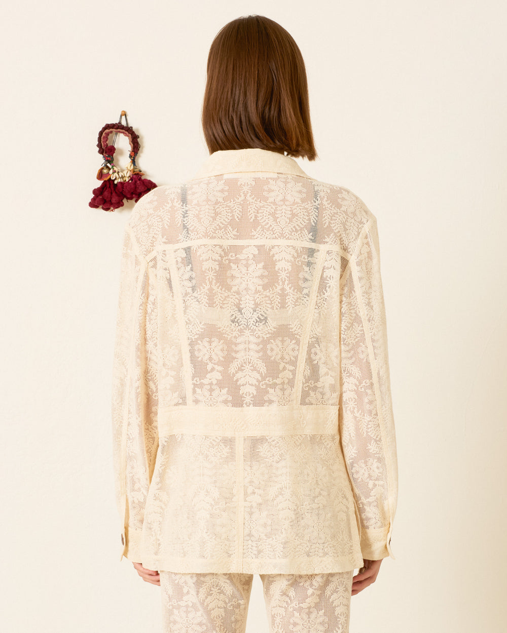 A person with shoulder-length brown hair stands with their back to the camera, wearing a sheer embroidered cream blouse and pants, topped with the Reno Pearl Lace Lattice Jacket, against a plain wall featuring a small hanging.