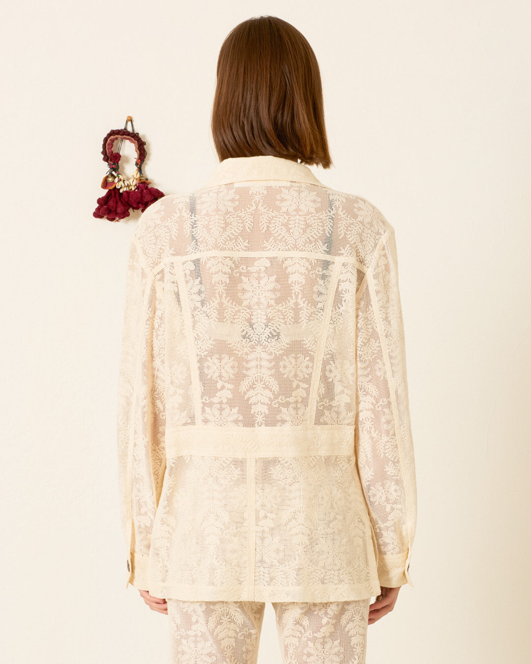A person with shoulder-length brown hair stands with their back to the camera, wearing a sheer embroidered cream blouse and pants, topped with the Reno Pearl Lace Lattice Jacket, against a plain wall featuring a small hanging.