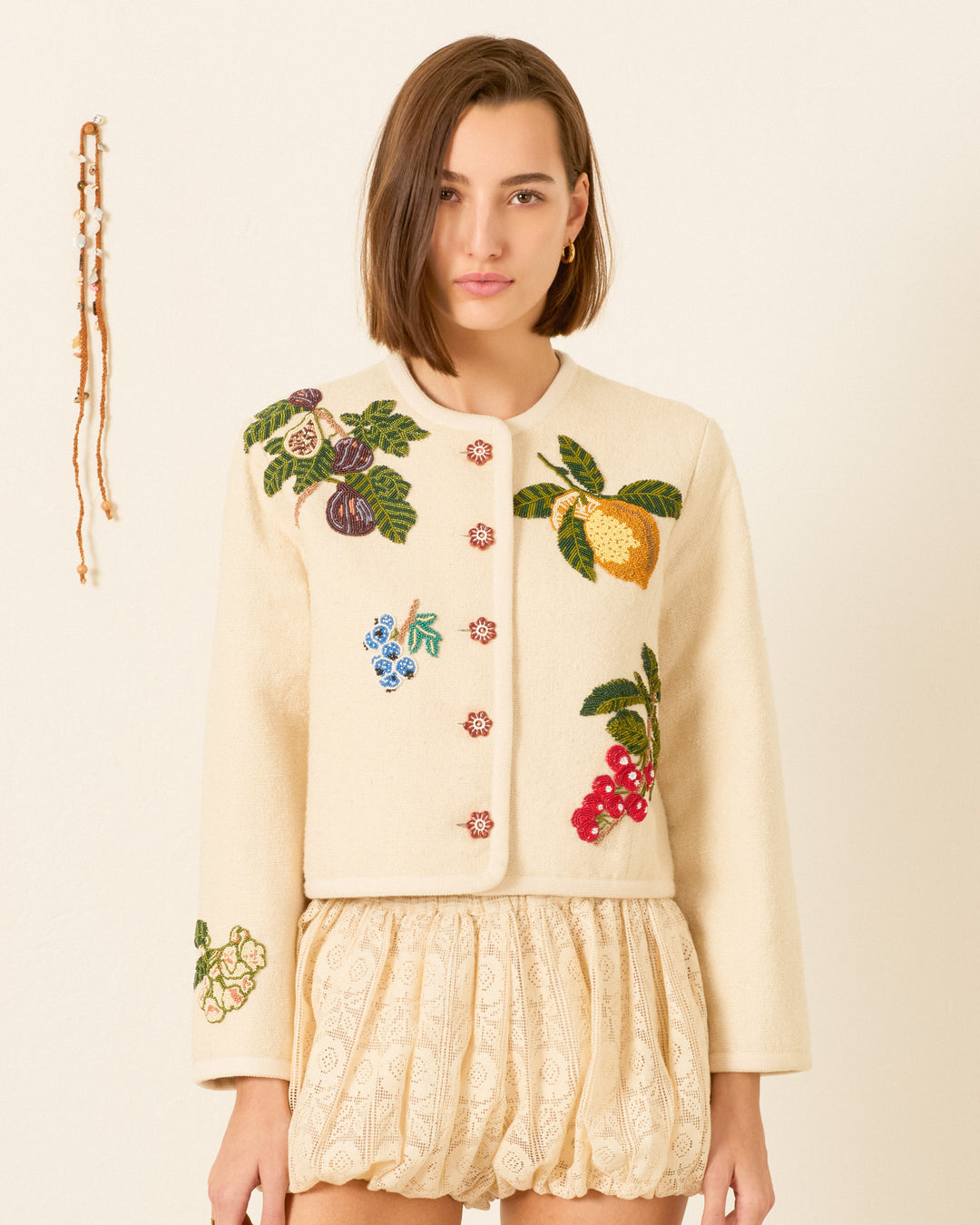 A woman wears the Rowan Nine Fruit Bead Jacket—a cream khadi cotton jacket featuring an Italian summer fruit motif—with a cream patterned skirt, standing before a plain backdrop and a beaded wall hanging.