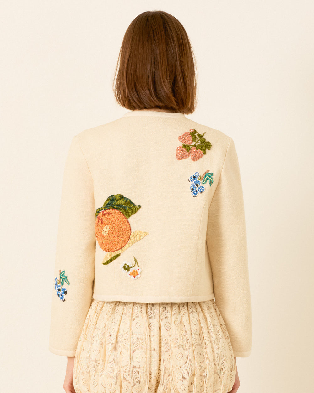 A woman facing away wears the Rowan Nine Fruit Bead Jacket—cream, hand-beaded with Italian summer fruit and floral embroidery—paired with a cream lace skirt.