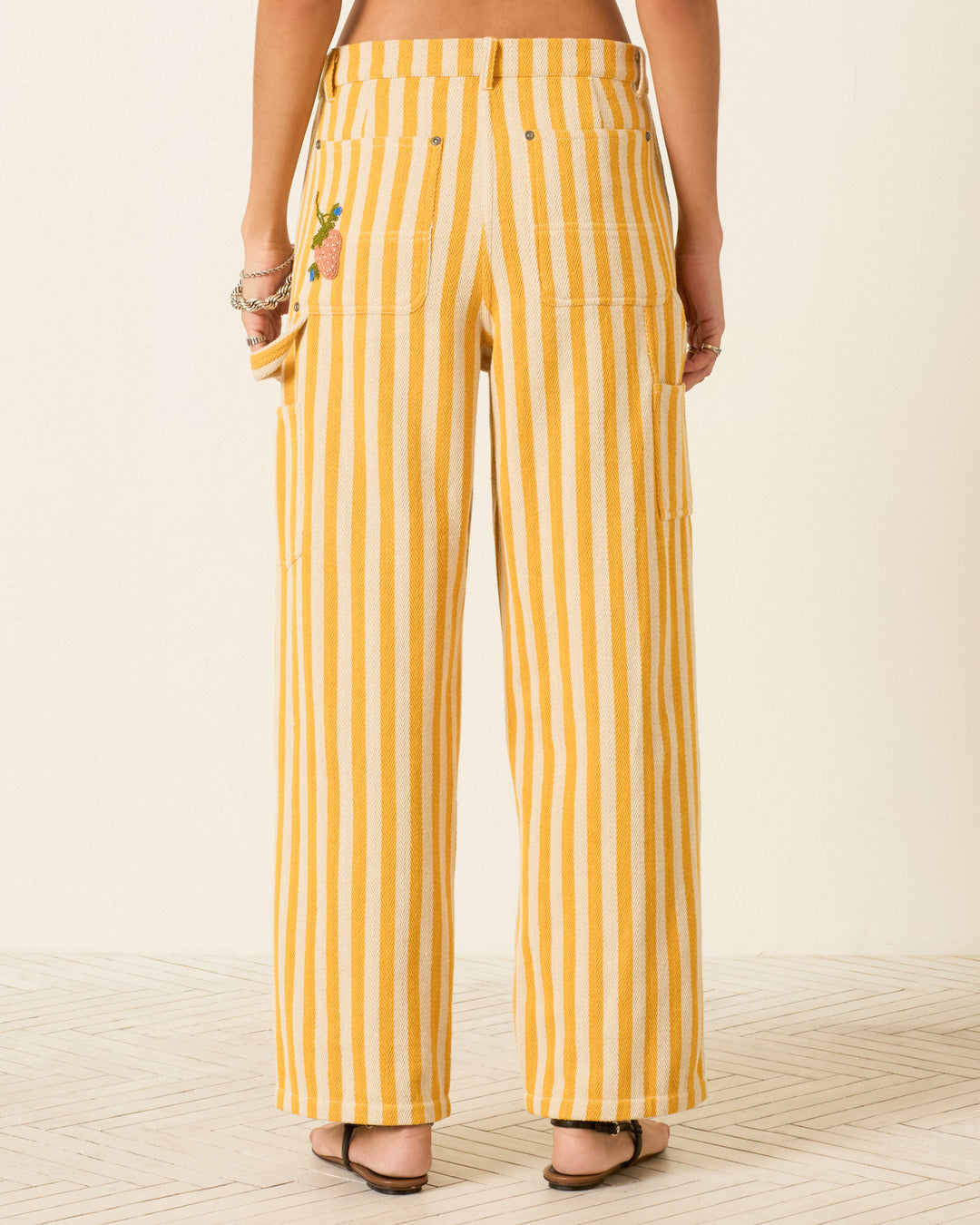 Someone stands on a light patterned floor wearing the Scottie Sunlight Stripe Pant, a high-rise straight-leg cotton twill pant with yellow and white vertical stripes, back pockets, and a small pineapple embroidery.