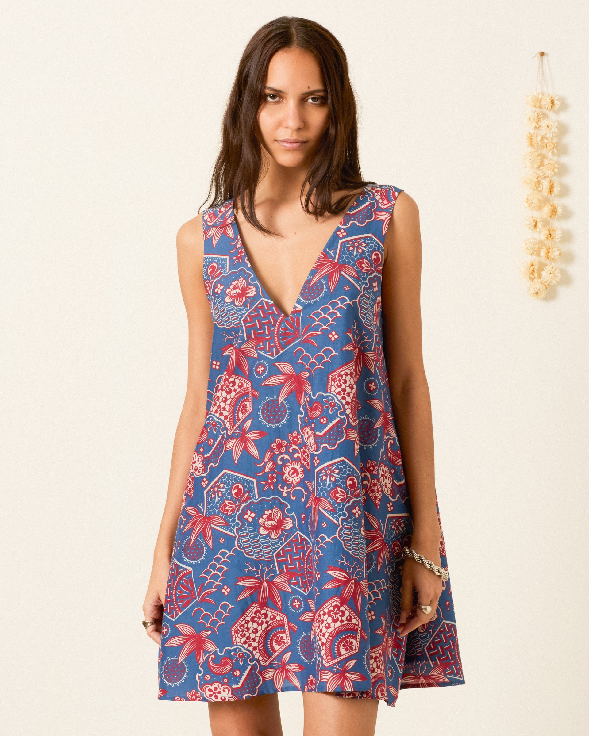 A woman stands against a plain wall, wearing the Soleil Delft Hive Dress—a sleeveless cotton-silk trapeze dress featuring a deep V-neck and a blue and red floral pattern. A small garland hangs on the wall beside her.