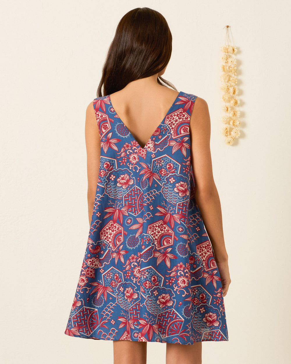 A woman stands against a plain wall with a hanging beige decoration, wearing the Soleil Delft Hive Dress—a sleeveless trapeze silhouette featuring blue and red wagara print and a V-shaped back.