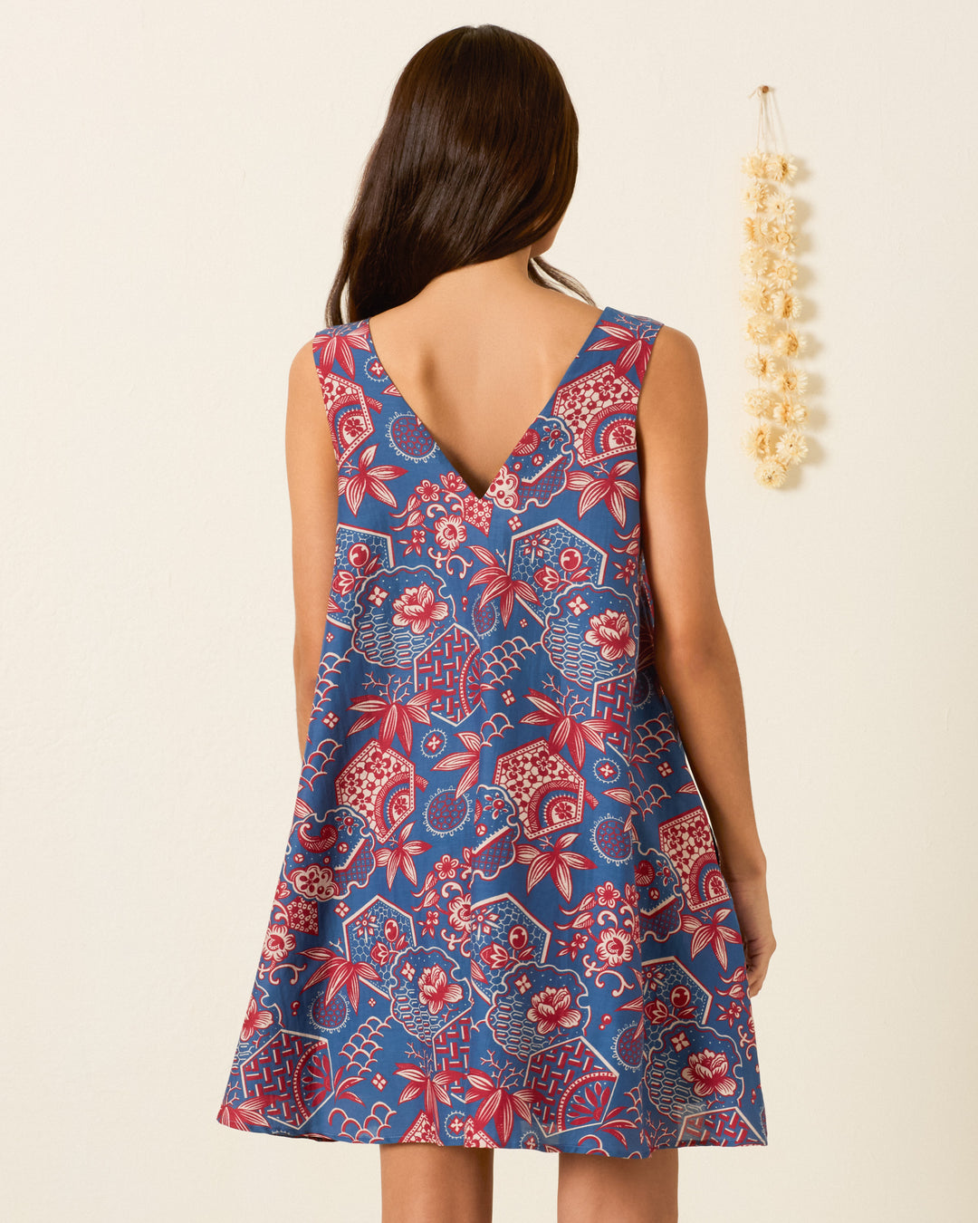 A woman stands against a plain wall with a hanging beige decoration, wearing the Soleil Delft Hive Dress—a sleeveless trapeze silhouette featuring blue and red wagara print and a V-shaped back.