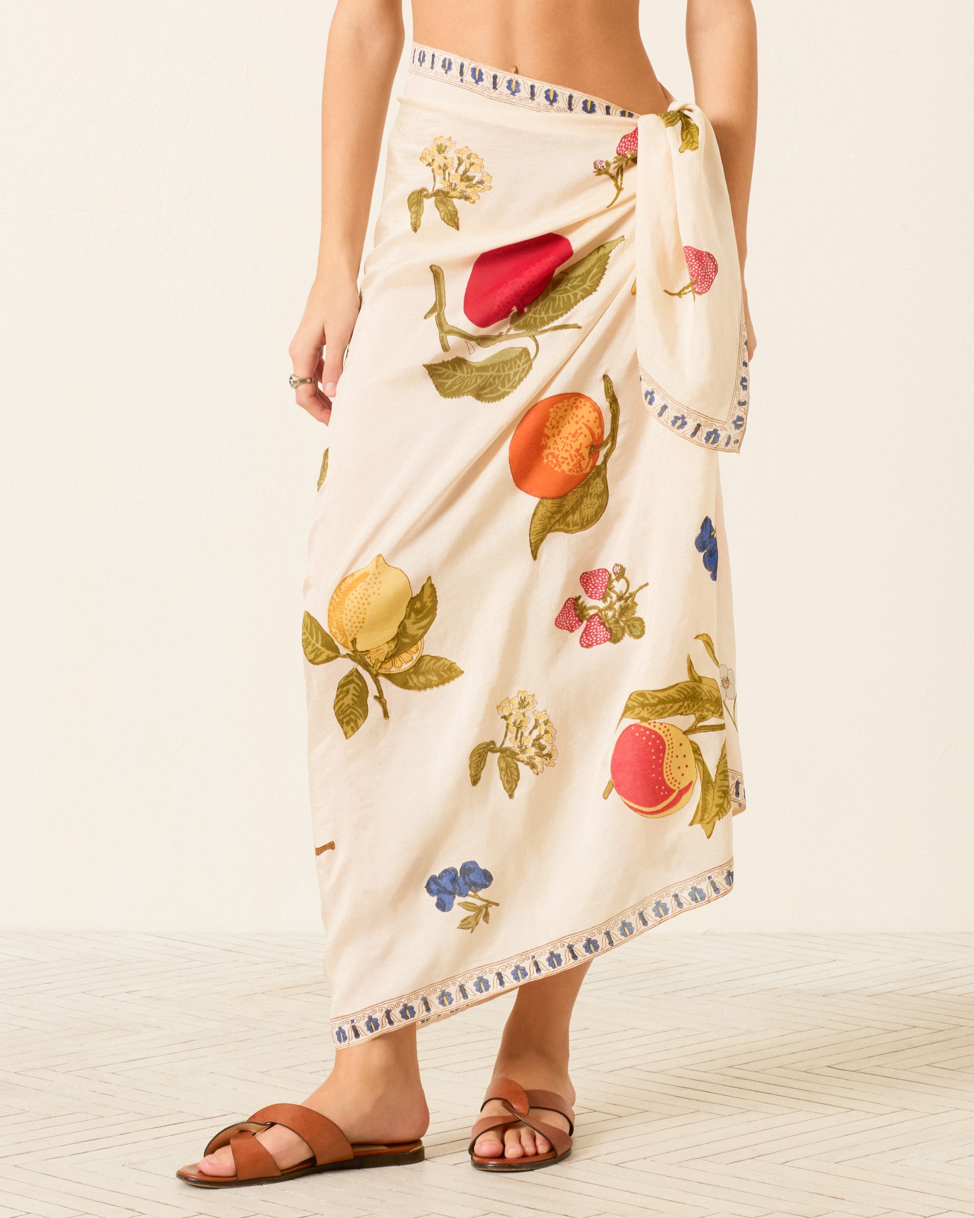 Wearing the Soraya Nine Fruit Silk Sarong, featuring a cream hand-block-printed Italian summer fruit motif, and brown sandals, a person stands on a light patterned floor.