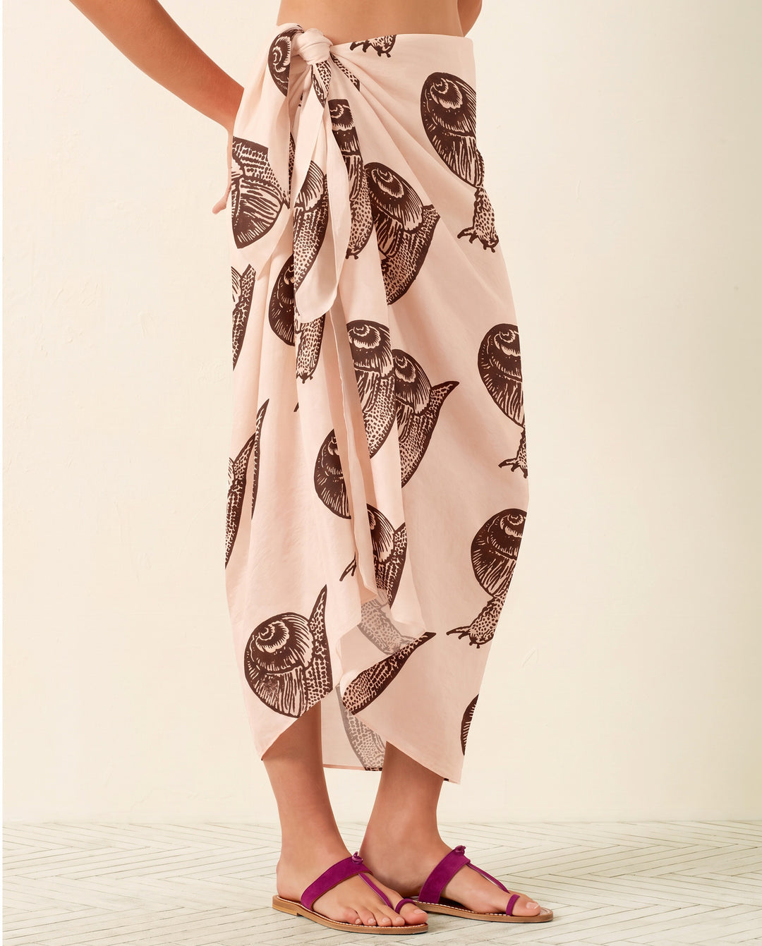 Wearing the Soraya Snail Study Silk Sarong, a light pink hand-block-printed piece, paired with purple sandals, this person stands on a light patterned floor against a plain background.
